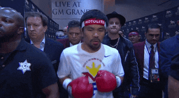 Sports gif. Boxer Manny Pacquiao walks out from the locker room surrounded by his posse. He's wearing boxing gloves and a white t-shirt, boxing the air slightly as he walks out. He's ready to rumble.