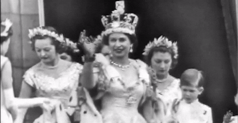 A black and white video clip of Queen Elizabeth in the 50's wearing her crown and cape and waving. She is surrounded by youn women with laurel wreaths in their hair.