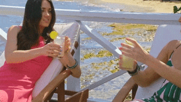 Reality TV gif. Two women from the show Coupled lounge on beach chaises and clink glasses.