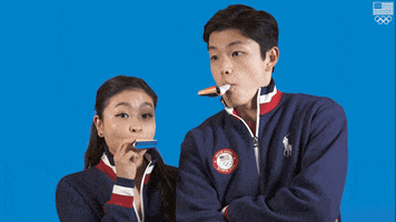 Sports gif. Alex and Maia Shibutani, figure skaters, blow brightly colored party horns over and over with neutral yet focused expressions against a blue backdrop. An American flag and Olympics logo appear in the top corner. 