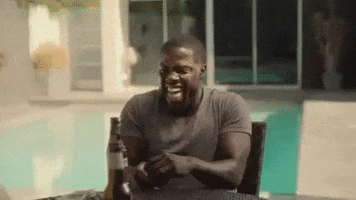 TV gif. Kevin Hart on Real Husbands of Hollywood sits at a table poolside with a beer in front of him. He chuckles and bounces his shoulders up and down with each laugh. 