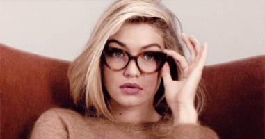 Celebrity gif. Seated on a brownish-orange couch and wearing a fuzzy beige sweater, Gigi Hadid tilts her glasses to give us a look of surprise.