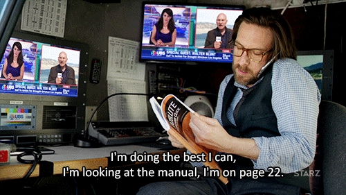 Gif of a man sitting at a desk surrounded by monitors. He's talking on the phone while leafing through a book, saying: "I'm doing the best I can, I'm looking at the manual, I'm on page 22." 