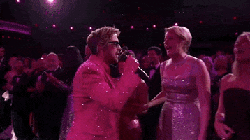 Oscars 2024 GIF. Ryan Gosling, dressed in a Barbie pink tuxedo and sunglasses, puts a microphone in the faces of Greta Gerwig, Margot Robbie, and America Ferrera, who enthusiastically sing, “I’m Just Ken!”