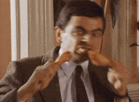 TV gif. Holding a fried chicken leg in each hand, a hungry Rowan Atkinson as Mr. Bean takes bites of chicken, bouncing back and forth from one hand to another.