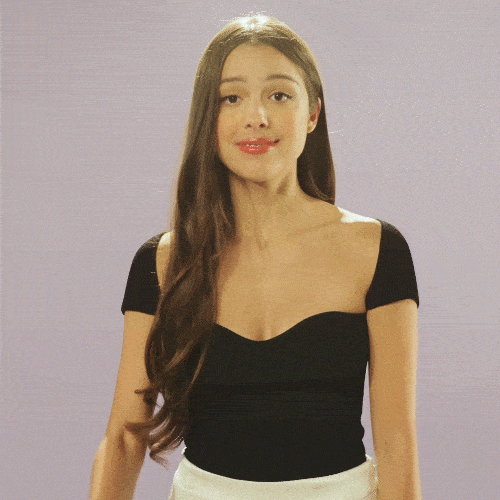 Video gif. Olivia Rodrigo smiles and gives a big wave at us against a white background.