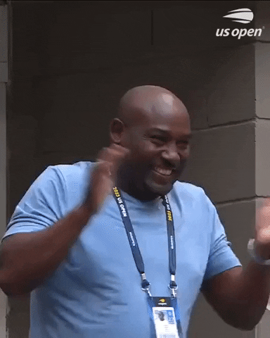 Video gif. Corey Gauff, father of Coco Gauf, grins from ear to ear as he claps his hands and points both fingers at someone offscreen like he's overcome with excitement, wearing a US Open badge. 