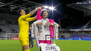 Sports gif. Kailen Sheridan and Kyra Carusa from the San Diego Wave in the NWSL are on the field before or after a game. Kyra holds out her arms with a bunch of team swag draped all over her like she's the display. Kailin tops it all off by putting another hot pink shirt or shorts over Kyra's head, patting her on the shoulder as she walks away. 