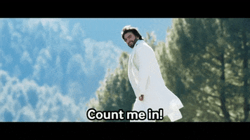 Movie gif. Ranveer Singh as Rocky in "Rocky Aur Rani Ki Prem Kahani" wears a white jacket and white scarf as he smiles and confidently walks across a forest background, shoulder-length hair blowing in the breeze and looking suave. Text, "Count me in!'