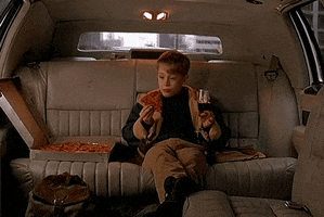 Movie gif. Macaulay Culkin as Kevin in Home Alone 2: Lost in New York, sits in the back seat of a limo with his legs crossed, holding a wine glass with juice in it, and eating a slice of pizza like he's living that rich life.