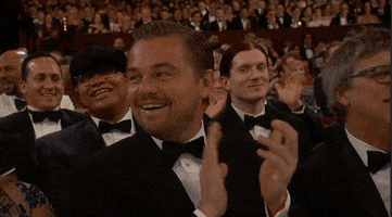 Celebrity gif. Leonardo DiCaprio sits in the audience of the Academy Awards. He looks over his shoulder with a large smile and claps along with the crowd. 