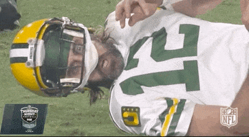 Sports gif. Aaron Rodgers of the Packers lies on the ground on his side with his helmet halfway on his head, staring out and appearing shocked.