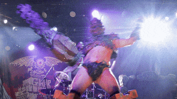 Celebrity gif. Member of GWAR dances on a stage with bright flashing lights, wielding a chainsaw, wearing blocky machine-like boots, and a jockstrap.