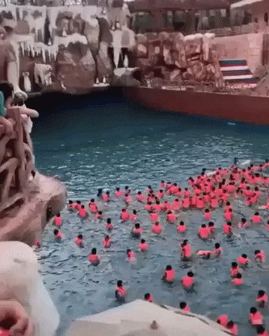Video gif. A bunch of people wearing pink life vests are in a wave pool and they all eagerly wait for the wave to come. A huge wave approaches the crowd and slams down, pushing everyone further back in the pool.