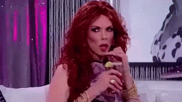 Reality TV gif. A bunch of drag queens from RuPaul's Drag Race are sitting together sipping drinks from straws while they eyeball one another. Everyone looks like they're waiting for someone to say something first, as they sassily sip their drinks.