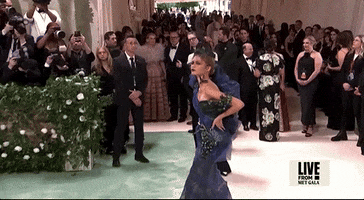 Met Gala 2024 gif. Zendaya poses on the red carpet wearing a midnight blue and dark teal dress with a large tulle puffed sleeve over her right shoulder and a net mesh and tulle feathered fascinator. She reaches up to touch an abstract bird detailing resting at the base of her bare neck. 