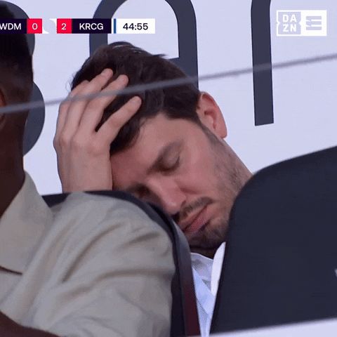 Video gif. Man sits in a stadium at a soccer game, alternating between putting his head in his hand and watching the game with a worried and defeated expression. 