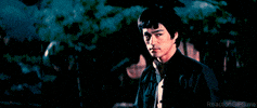Movie gif. Bruce Lee as Chen Zhen in Fist of Fury scowls, determined, as he rips off his shirt and throws it to the ground.
