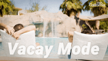 Sponsored gif. Two people relax in lounge chairs that sit in the shallow water of a luxurious pool in California. On the other side of the pool, a small waterfall cascades from a large rock formation. Text reads, “Vacay mode.”