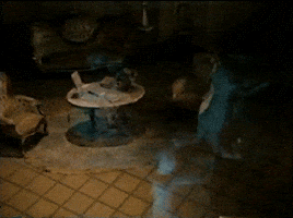 Video gif. Three holographic dancing ghost couples from the Haunted Mansion ride at Disneyland dance in circles around a dark, elegant living space. 