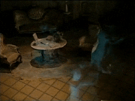 Video gif. Three holographic dancing ghost couples from the Haunted Mansion ride at Disneyland dance in circles around a dark, elegant living space. 