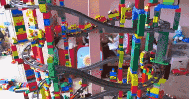 Video gif. On an intricately built but fragile lego tower, a toy train moves up a track for a moment, then the entire structure crumbles to the floor.
