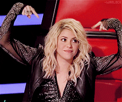 TV gif. Musician Shakira raises her hands over her head and points to herself from her judge's seat on The Voice. 