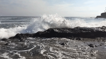 Video gif. Ocean waves crashing onto a large rock on Laguna beach.