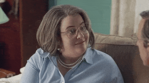 Women wearing glasses and blue shirt in conversation with man on sofa. Woman turns to camera, winks and smiles.