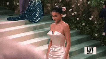 Met Gala 2024 gif. Slow motion close up of the top of Kylie Jenner's Oscar de la Renta champagne-colored strapless column gown featuring a pointed bust. She also wears a pale pink rose in a smooth mid bun.