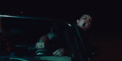 Music video gif. Yungblud in the music fideo for The Funeral hangs his hand out of a car window, screaming wildly, as he holds onto the steering wheel.