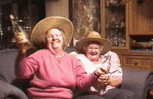 Video gif. Two elderly women sit on a couch with bottles of liquor and dance as they drink. 