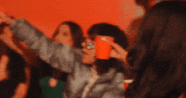 Music video gif. From the video for La Diabla by Xiao, a group of women dressed for a nightclub clink red solo cups together in a room cast in red light, looking cool and happy, like they're having the best night out. 