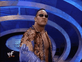 Sports gif. Dwayne The Rock Johnson on WWE Smackdown snaps his arm up and holds his hand out signaling to stop. He wears blacked out sunglasses and a stone cold look on his face.
