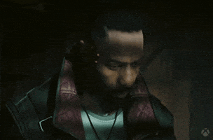 Video game gif. In "Cyberpunk 2077," a man wearing a leather jacket looks off with a pensive expression, shadows casting across his face in a dark room. 