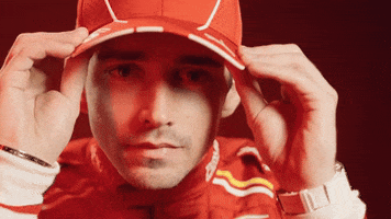 Celebrity gif. Charles Leclerc, a Formula 1 driver, wears a red racing suit and a red baseball hat. He looks at us with a serious expression on his face as he adjusts his hat and bats the camera way with his hand. 