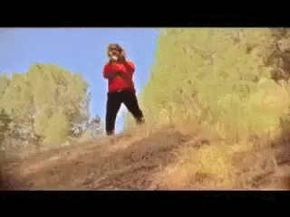 Gif of a man with shaggy hair and sunglasses wearing a red sweatshirt playing the trumpet on top of a hill and doing a jiggly dance .