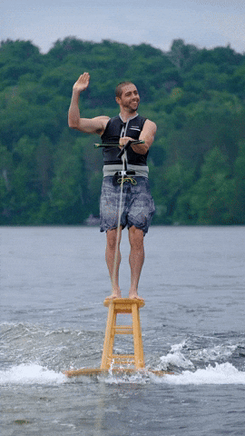 Video gif. A smiling man wakeboarding on a barstool attached to a tabletop waves his hand in a cupping motion, like the queen.