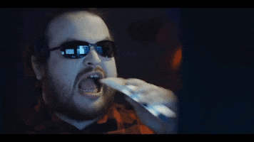 Music video gif. From Amon Amarth video for "Keyboard Warrior," man wearing sunglasses inside, looking at a screen while chomping on a hot pocket.