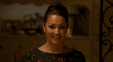 TV gif. Paula Garcés as Geny on On My Block. She's sitting at a dining table with a stiff smile on her face as she looks around awkwardly.