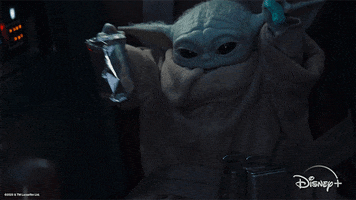 Star Wars gif. Baby Yoda bobs in a seat of the Mandalorian's cruiser, green hands swaying excitedly above his large pointed ears. He has wide, dark eyes and an open mouth, holding tight to a cookie in his hand. 