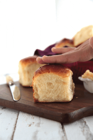A white bread roll on a dark brown wooden board, being patted down by a caucasian coloured hand.