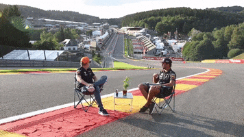 Sports gif. Daniel Ricciardo and Max Verstappen sit in lawn chairs on an F1 race track. They spring up from their chairs, elated, and immediately begin dancing, throwing out the silliest moves they can fathom.