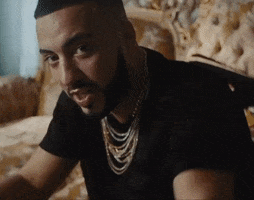 Music video gif. French Montana raises a low-ball glass with brown liquor as he smiles and winks towards us.