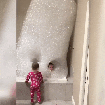 Video gif. Four children in a bathroom with two in the tub surrounded by a tower of bubbles all the way up to the ceiling. One of the kids appears inside a hole in the bubbles and jumps with a smile on his face. 