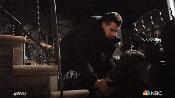 TV gif. Octavio Pisano as Joe on Law & Order : Special Victims Unit, leans over an unconscious woman, picking her up from her face down position at the foot of a staircase. 