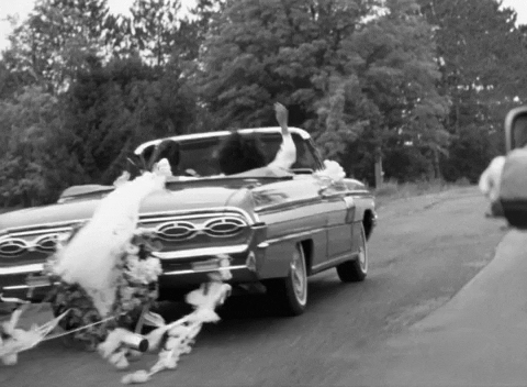 Černobílý obrázek s ujíždějícím autem se svatebním párem a zavěšenými plechovkami na zadním nárazníku. 