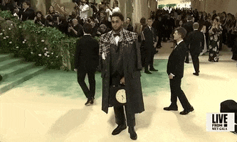Met Gala 2024 gif. Ben Simmons poses on the red carpet wearing a Thom Browne black leather suit and jacket with black, white, and red plaid detailing around the shoulders. He's holding a black rectangular briefcase with a cream-colored clock face on the front. 