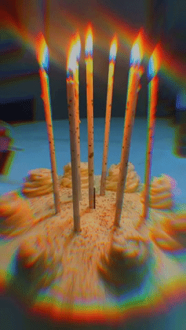 Video gif. Closeup of a birthday cake with tall candles, casting trippy beams of rainbow light across the frame. 
