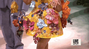 Met Gala 2024 gif. Closeup of the top of Nicki Minaj's yellow Marni mini dress with a low-cut v-neckline and 3D multicolored metal flowers that seem to sprout all around her. The dress flares out in a stiff structured pattern from her waist and comes to about her mid thigh.
